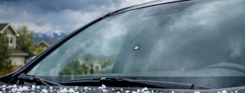 Close-up stock photo of a car windshield with a chip and hailstones on the hood, outdoors in a Colorado suburb with stormy spring sky.